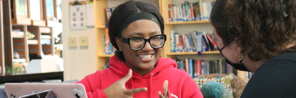 A student smiles and animatedly talks to an 826 Boston team member. 