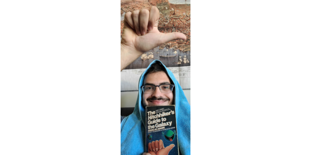 Mark holds up the hitchhikers gesture while holding a copy of the Hitchhiker's Guide to the Galaxy