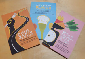 On a table are three student authored publications. From left to right, "A Long Walk to Healthy," "All Kinds of Flavor," and "With A Crunch and A Slurp."