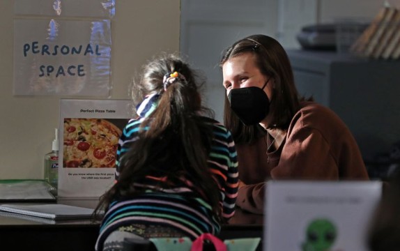 A student with long hair and a colorful sweatshirt faces a tutor with short brown hair and a mask.