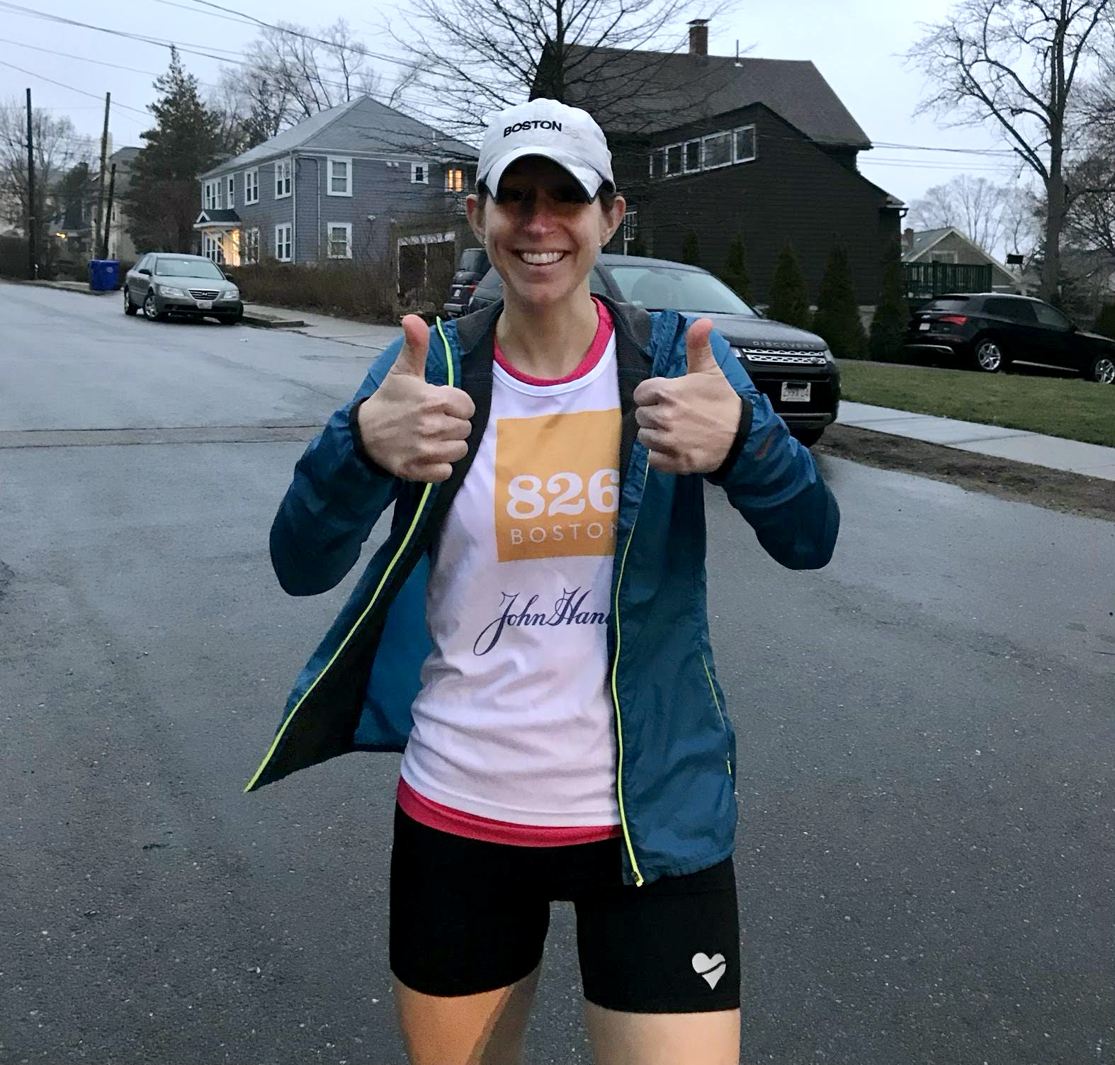 Erin Sunderland stands outside wearing a hat and a 826 boston white tshirt and running shorts. She is posing with two thumbs up