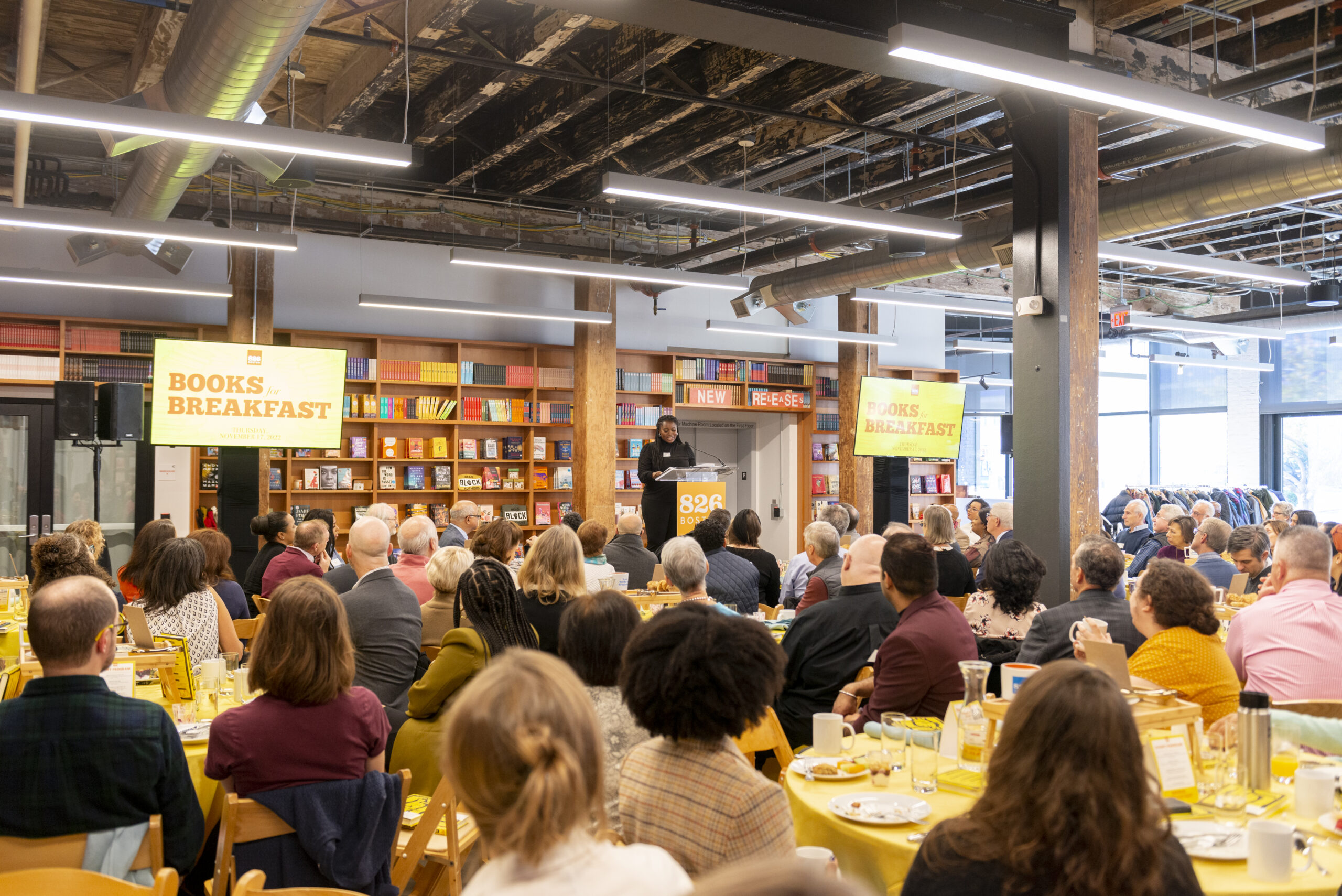 A wideshot of Books for Breakfast fundraiser. The audience is seated and is listening to a speaker who is standing on a podium. 
